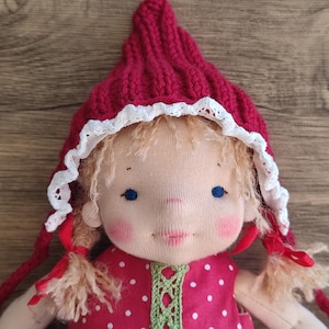 May include: A handmade doll with a red knitted cap trimmed with white lace. The doll has a red polka-dot dress with green trim, blonde hair, and rosy cheeks. The doll's eyes are blue.