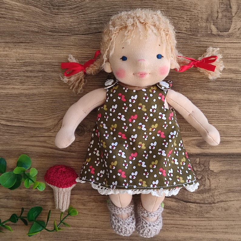 May include: A handmade cloth doll with blonde pigtails tied with red ribbons. The doll wears a green floral dress with white lace trim and gray crocheted shoes. A small, red mushroom decoration sits nearby.
