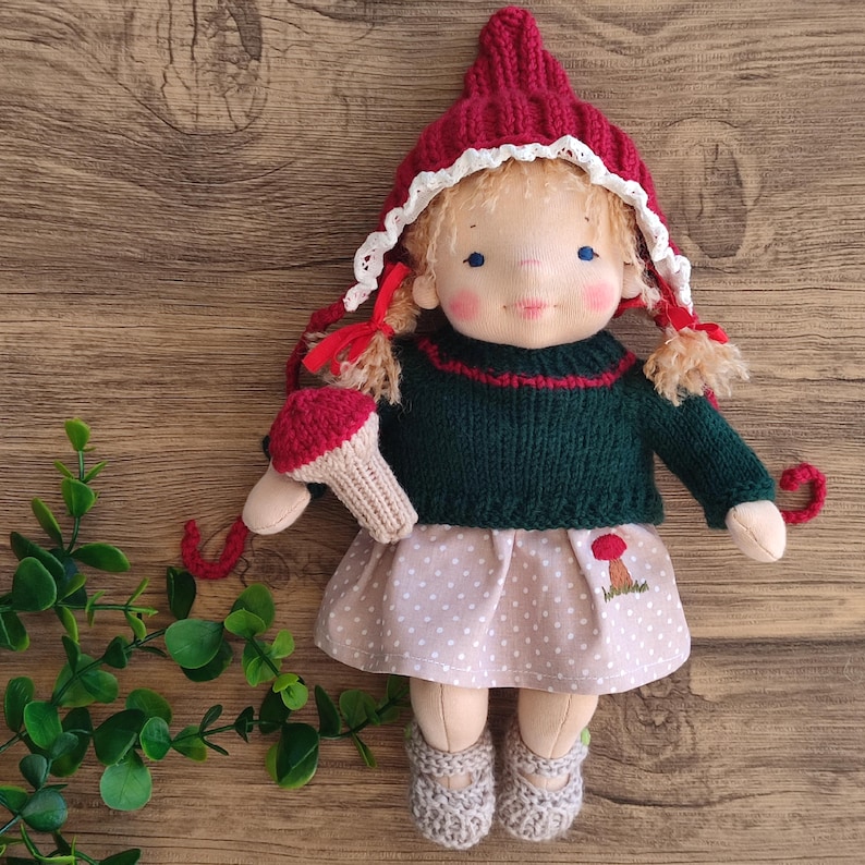May include: A handmade doll with blonde pigtails, wearing a red knit hat with white lace trim, a green sweater, and a polka-dot skirt. The doll holds a mushroom and wears gray knit shoes. A mushroom is embroidered on the skirt.