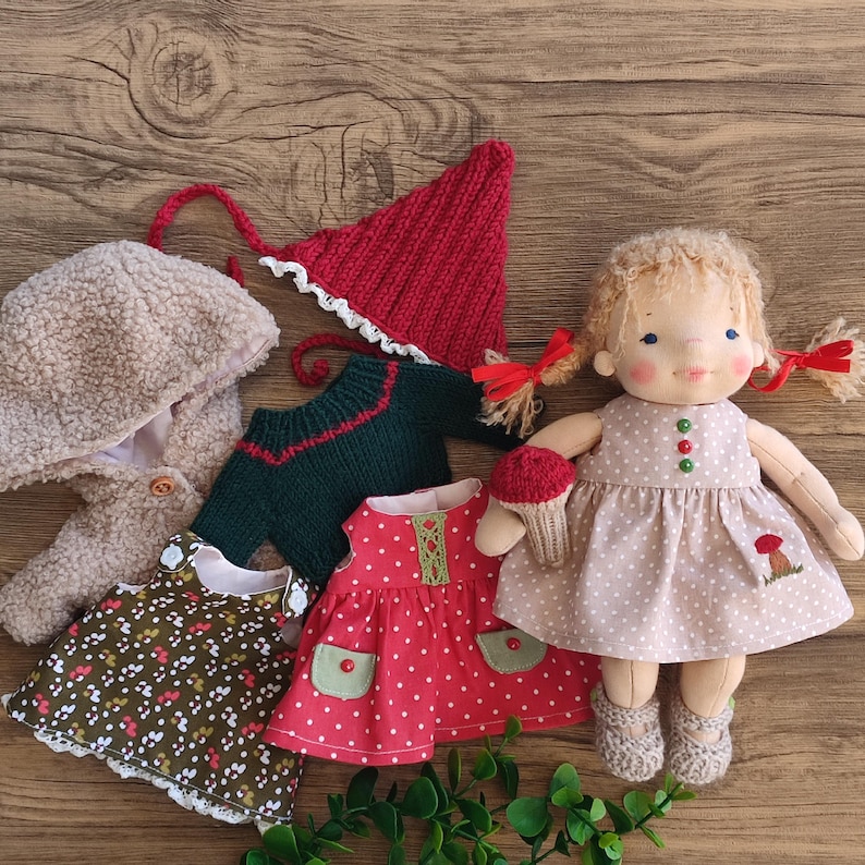 May include: A handmade doll with blonde pigtails wearing a polka dot dress, surrounded by miniature clothing items. The clothing includes a red knit hat, a green sweater, and two dresses. The doll is holding a small cupcake.