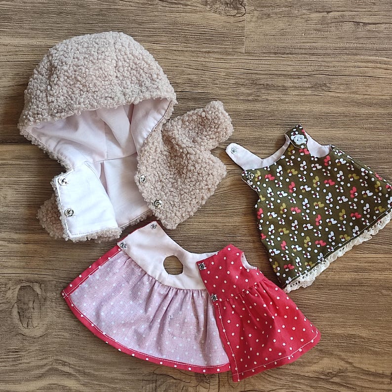 May include: Three pieces of doll clothing: a beige hooded jacket, a red polka dot dress, and a green floral dress. The jacket has a soft, textured exterior and a white lining. The dresses have contrasting trim and are designed for a doll.