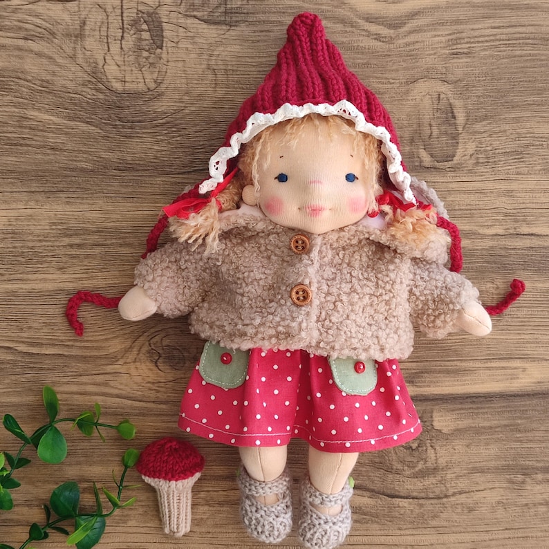 May include: A handmade doll wearing a red knit cap with white lace trim, a tan fuzzy jacket, and a red polka dot dress. The doll has blonde hair, rosy cheeks, and gray shoes. A small knitted mushroom is next to the doll.
