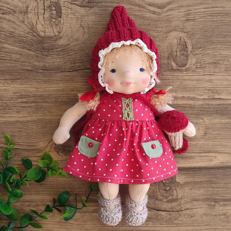 May include: A handmade doll dressed in a red polka-dot dress with green pockets and a matching knitted hood. The doll has a painted face, blonde hair, and gray shoes. It holds a red mushroom.