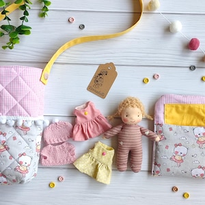 Op de afbeelding: Een handgemaakte pop met blond haar en gestreepte kleding is omringd door miniatuurjurkjes en gebreide tops in roze en geel. Twee stoffen tassen met een roze ruitpatroon en een berenprint zijn ook aanwezig. Het label luidt "Tanitadolls".