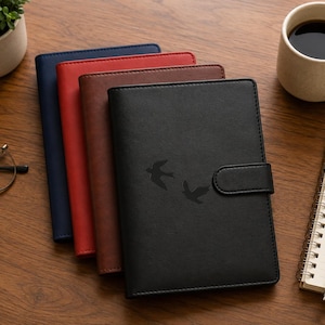 May include: A stack of four rectangular notebooks with textured covers in black, brown, red, and navy blue. The black notebook features a magnetic closure tab and a debossed graphic of two flying birds on the front cover, with a smooth finish.