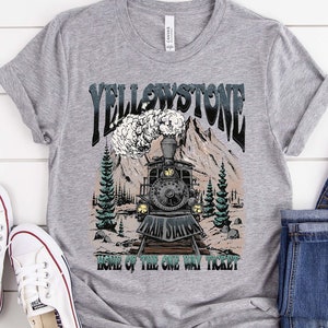 Op de afbeelding: Een grijs t-shirt met een afbeelding van een vintage trein voor bergen en bomen, met het woord "YELLOWSTONE" erboven en "HOME OF THE ONE WAY TICKET" eronder. Het shirt wordt gecombineerd met een blauwe jeans en witte sneakers.