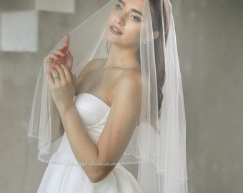 May include: A bride wearing a white strapless wedding dress with a long, white veil. The veil has a beaded trim and is draped over the bride's head.