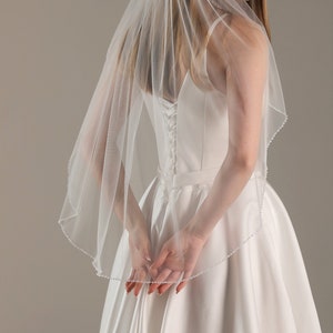 May include: A sheer white bridal veil with a beaded trim. The veil is draped over the shoulders. The bride is wearing a white satin wedding dress with a corset back.