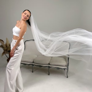May include: A woman wearing a white tube top and high-waisted white pants, with a long, flowing white veil. The veil is billowing in the air. A vintage-style sofa is in the background.