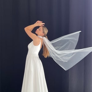 May include: A white bridal gown with a flowing skirt and a sleeveless bodice. A sheer, white veil adorned with small, scattered pearls is attached at the back. The veil is flowing in the wind. The background is a solid black.
