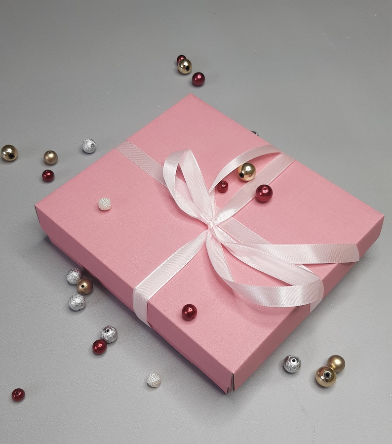 Puede incluir: Una caja de regalo rosa con una cinta de sat&eacute;n blanca atada en un lazo. La caja est&aacute; rodeada de peque&ntilde;as cuentas de colores.