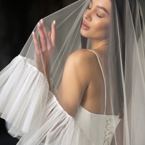 Puede incluir: Una novia con un vestido de novia blanco con la espalda de encaje y un velo de tul blanco. El velo tiene un borde con volantes y est&aacute; drapeado sobre la cabeza de la novia.