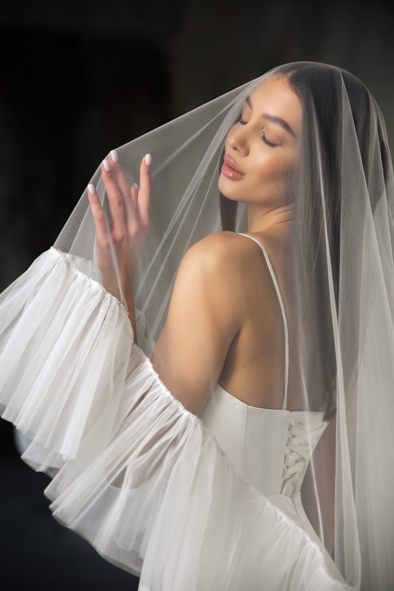 Puede incluir: Una novia con un vestido de novia blanco con la espalda de encaje y un velo de tul blanco. El velo tiene un borde con volantes y est&aacute; drapeado sobre la cabeza de la novia.