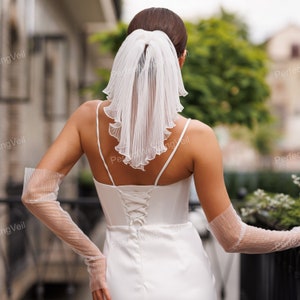 Peut inclure: Une mariée porte une robe de mariée blanche avec un dos en corset et des bretelles fines. Elle porte un voile blanc plissé et de longs gants transparents avec des détails scintillants. L'image est prise de dos, mettant en valeur la tenue de mariée élégante.