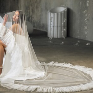 Puede incluir: Un velo de novia blanco con un borde con volantes, usado por una persona sentada en el suelo. El velo est&aacute; hecho de una tela transparente y tiene una cola larga.