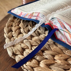 May include: An open book with a dark blue suede cover, featuring white lace trim along the page edges and a solid dark blue ribbon bookmark. The page edges display a colorful floral pattern, and the spine includes a thin red accent ribbon.
