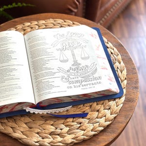 May include: An open Bible with a blue cover and a white lace ribbon bookmark. The right page features a black and white line art illustration of a scale of justice with the text: The LORD will give justice to his people and have compassion on his servants. Psalm 135:14.