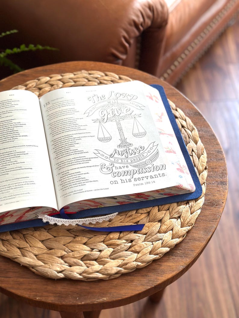 May include: An open Bible with a blue cover and a white lace ribbon bookmark. The right page features a black and white line art illustration of a scale of justice with the text: The LORD will give justice to his people and have compassion on his servants. Psalm 135:14.