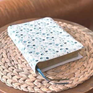 May include: A book with a cover featuring a light blue and green floral pattern. The book has a blue ribbon bookmark and is resting on a woven placemat. The book's pages have gold edges.