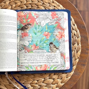 May include: An open book with a blue cover featuring a page decorated with watercolor-style floral illustrations, two small bird drawings, and the printed text Matthew 10:29-31, which reads about sparrows and the value of life in black ink.
