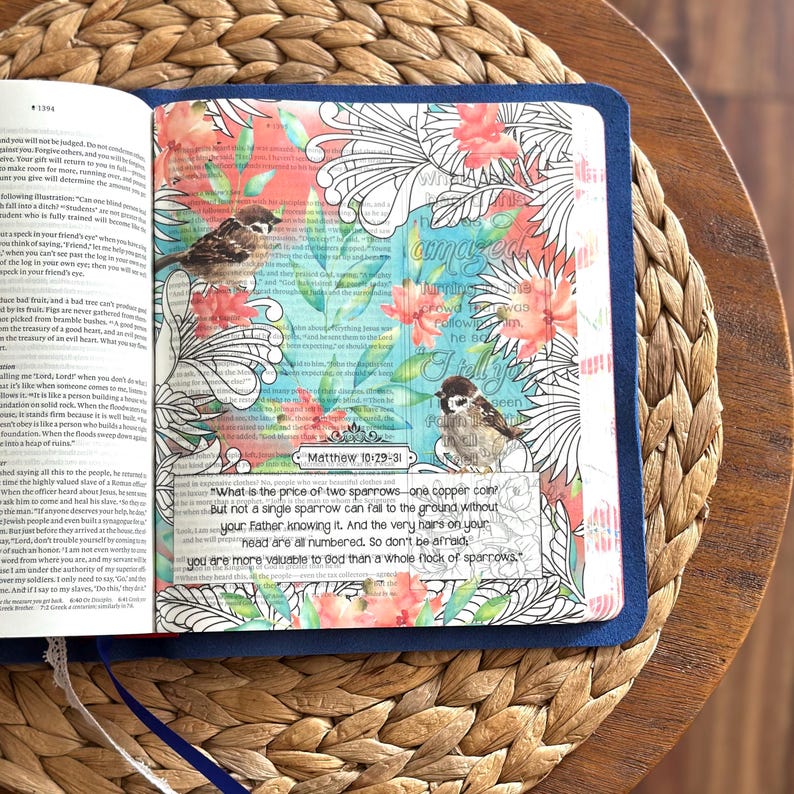 May include: An open book with a blue cover featuring a page decorated with watercolor-style floral illustrations, two small bird drawings, and the printed text Matthew 10:29-31, which reads about sparrows and the value of life in black ink.