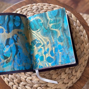 May include: An open journal with a dark purple cover and a white ribbon bookmark. The interior pages feature a marbled design with swirling patterns of metallic gold, deep teal, and bright blue paint, creating a textured and fluid visual effect.