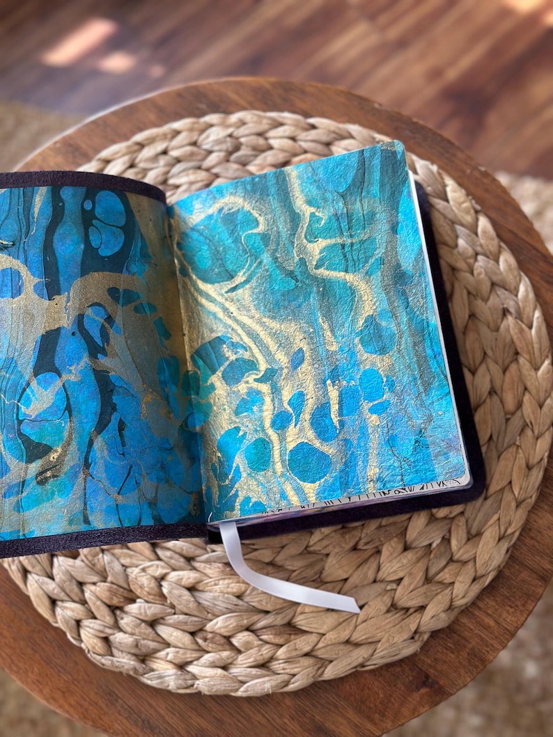 May include: An open journal with a dark purple cover and a white ribbon bookmark. The interior pages feature a marbled design with swirling patterns of metallic gold, deep teal, and bright blue paint, creating a textured and fluid visual effect.