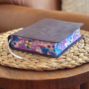 May include: A dark brown leather-bound book with a white ribbon bookmark. The page edges feature a vibrant, colorful print of blue and yellow butterflies, pink flowers, and intricate swirling patterns against a purple background.