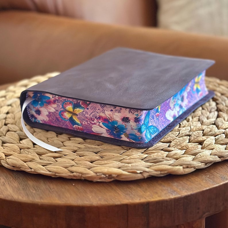May include: A dark brown leather-bound book with a white ribbon bookmark. The page edges feature a vibrant, colorful print of blue and yellow butterflies, pink flowers, and intricate swirling patterns against a purple background.
