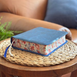 May include: A closed book with a solid matte blue cover and a floral pattern printed on the page edges. The floral design features red, orange, and green botanical illustrations. A thin blue ribbon bookmark with a white lace trim extends from the side.