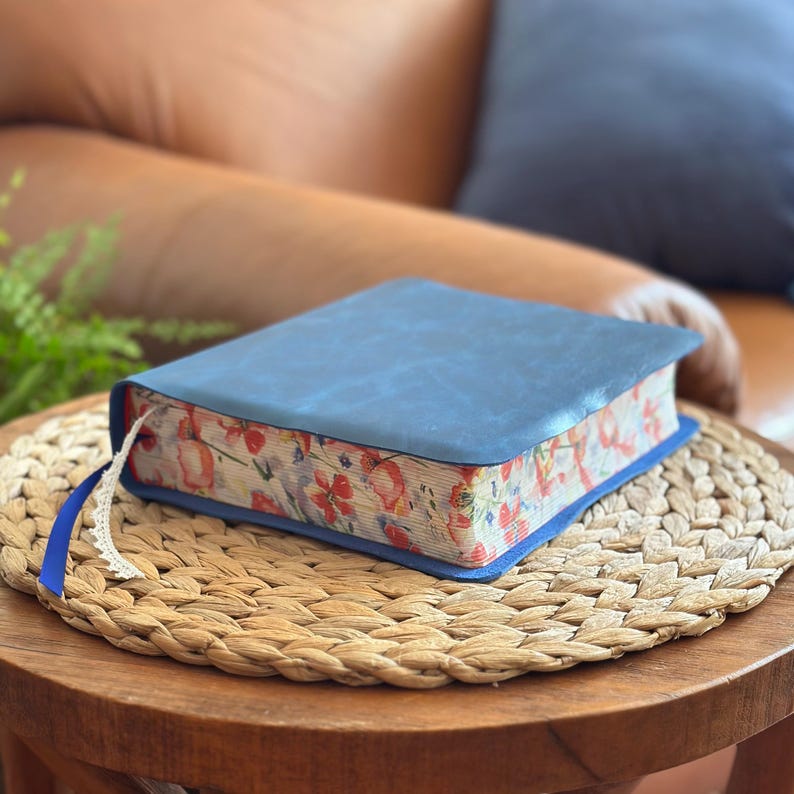 May include: A closed book with a solid matte blue cover and a floral pattern printed on the page edges. The floral design features red, orange, and green botanical illustrations. A thin blue ribbon bookmark with a white lace trim extends from the side.