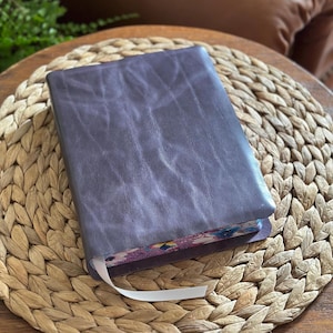 May include: A rectangular journal with a dark purple, textured leather cover. The edges of the pages feature a colorful floral pattern in shades of pink, blue, and green. A thin, white ribbon bookmark extends from the bottom of the book.