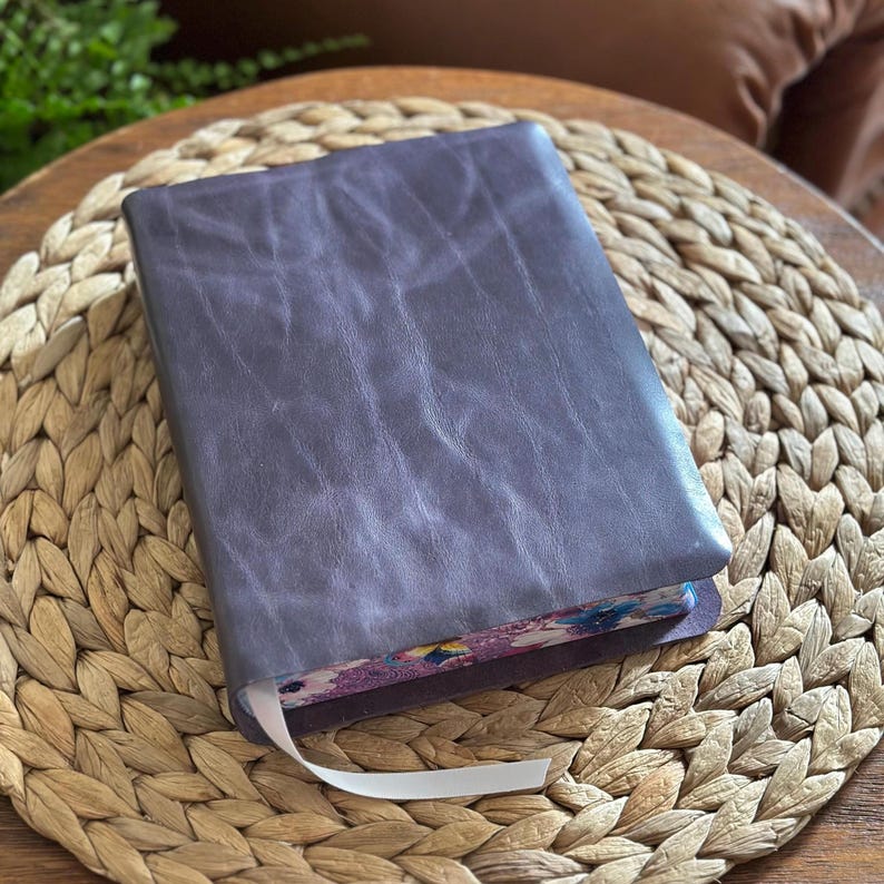 May include: A rectangular journal with a dark purple, textured leather cover. The edges of the pages feature a colorful floral pattern in shades of pink, blue, and green. A thin, white ribbon bookmark extends from the bottom of the book.