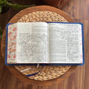 May include: An open Bible with a blue cover featuring intricate black and white line art illustrations and hand-lettered typography on the pages. The page edges are decorated with a colorful floral pattern, and a white lace ribbon bookmark is visible.