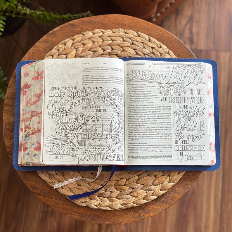May include: An open Bible with a blue cover featuring intricate black and white line art illustrations and hand-lettered typography on the pages. The page edges are decorated with a colorful floral pattern, and a white lace ribbon bookmark is visible.