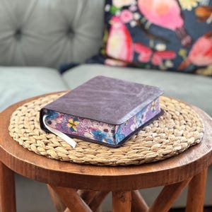 May include: A thick book with a dark brown, textured cover and a white ribbon bookmark. The page edges feature a vibrant, multi-colored floral and butterfly pattern in shades of purple, blue, pink, and yellow, visible along the side of the book.