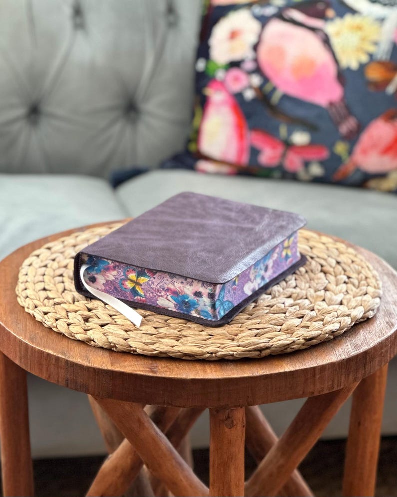May include: A thick book with a dark brown, textured cover and a white ribbon bookmark. The page edges feature a vibrant, multi-colored floral and butterfly pattern in shades of purple, blue, pink, and yellow, visible along the side of the book.