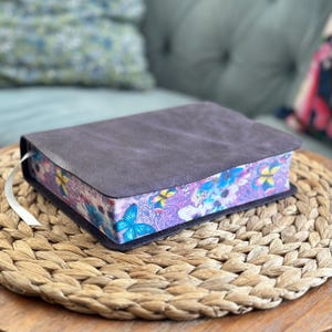 May include: A rectangular book with a dark gray, textured cover. The page edges feature a vibrant, colorful print of blue and yellow butterflies, purple paisley patterns, and various floral elements. A thin white ribbon bookmark extends from the side.
