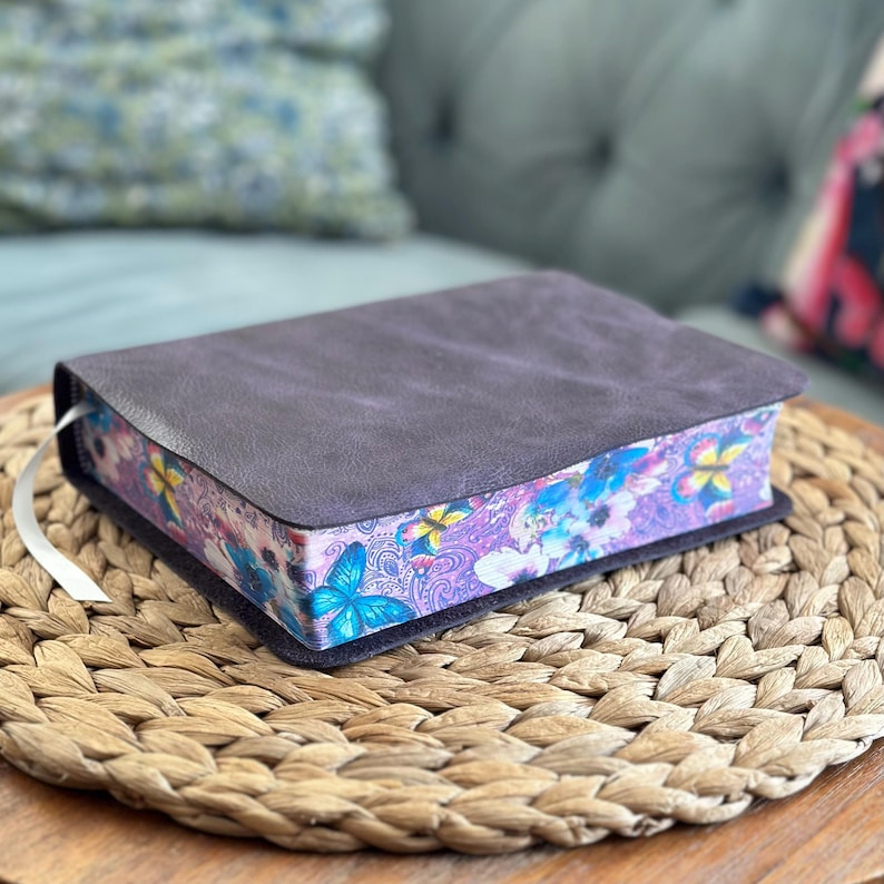 May include: A rectangular book with a dark gray, textured cover. The page edges feature a vibrant, colorful print of blue and yellow butterflies, purple paisley patterns, and various floral elements. A thin white ribbon bookmark extends from the side.