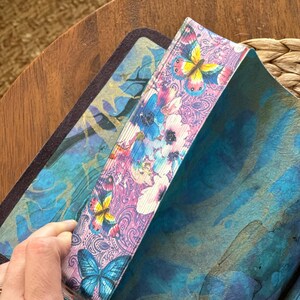 May include: A book with page edges decorated with a colorful print of butterflies and flowers in shades of pink, blue, and yellow. The interior pages feature a blue and teal watercolor-style wash with a dark, abstract pattern throughout.