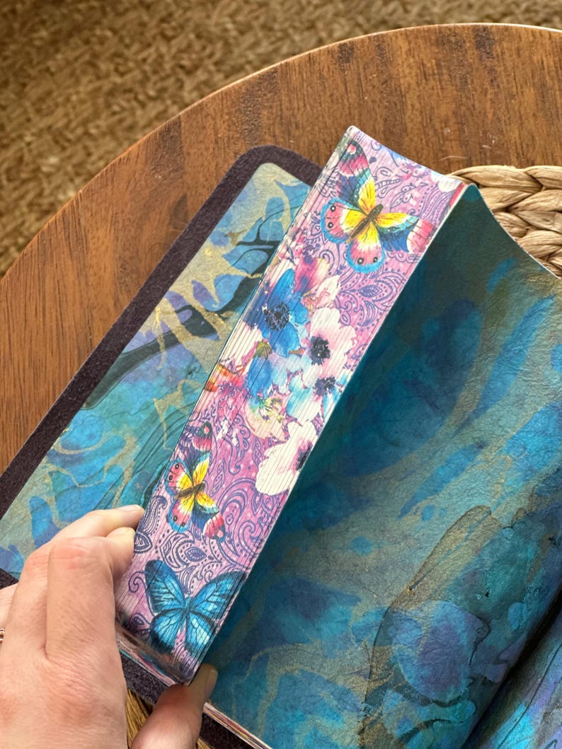 May include: A book with page edges decorated with a colorful print of butterflies and flowers in shades of pink, blue, and yellow. The interior pages feature a blue and teal watercolor-style wash with a dark, abstract pattern throughout.