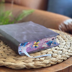 May include: A thick book with a matte dark gray cover and a white ribbon bookmark. The page edges feature a vibrant, colorful print of butterflies and floral patterns in shades of pink, blue, yellow, and purple against a detailed background.