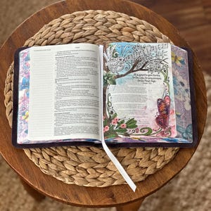 May include: An open Bible with a white ribbon bookmark. The left page contains text from 1 Chronicles. The right page features colorful illustrations of a bird, flowers, and a butterfly, with the text: It is good to give thanks to the Lord, to sing praises to the Most High. Psalm 92:1.