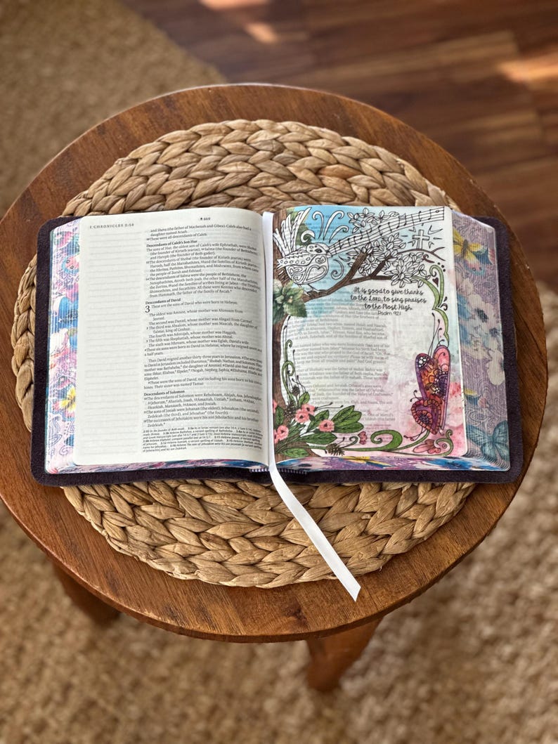 May include: An open Bible with a white ribbon bookmark. The left page contains text from 1 Chronicles. The right page features colorful illustrations of a bird, flowers, and a butterfly, with the text: It is good to give thanks to the Lord, to sing praises to the Most High. Psalm 92:1.