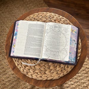 May include: An open book with dark covers and floral-patterned page edges. The left page contains printed text, while the right page features a circular line-art illustration of a helmet and the text: Believe in the Lord your God, and you will be able to stand firm; believe in his prophets, and you will succeed. 2 Chronicles 20:20.