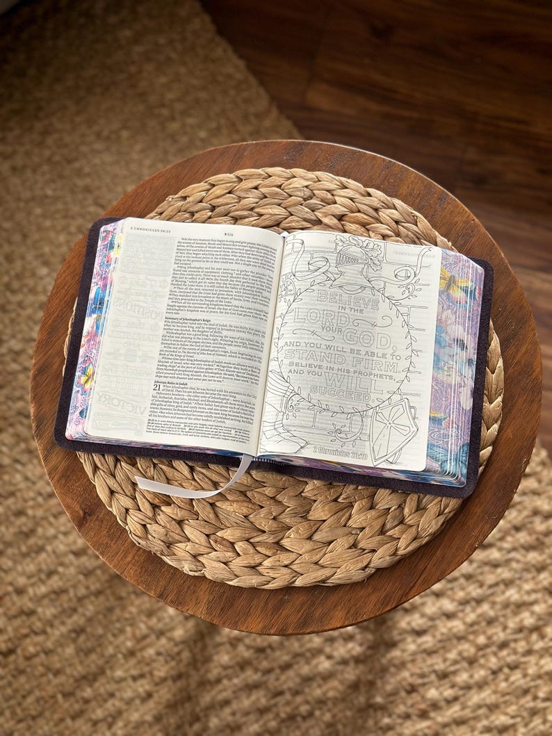 May include: An open book with dark covers and floral-patterned page edges. The left page contains printed text, while the right page features a circular line-art illustration of a helmet and the text: Believe in the Lord your God, and you will be able to stand firm; believe in his prophets, and you will succeed. 2 Chronicles 20:20.