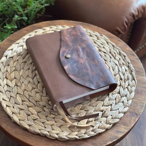 May include: A rectangular journal with a smooth brown leather cover and a textured, dark brown distressed leather flap closure secured by a single metal snap button. The journal includes two thin ribbon bookmarks in cream and dark brown colors.