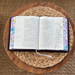 May include: An open Bible with dark covers and patterned page edges featuring butterflies and flowers. The right page displays a hand-drawn illustration of a hummingbird and a flower with the text, Sing a new song to the Lord!