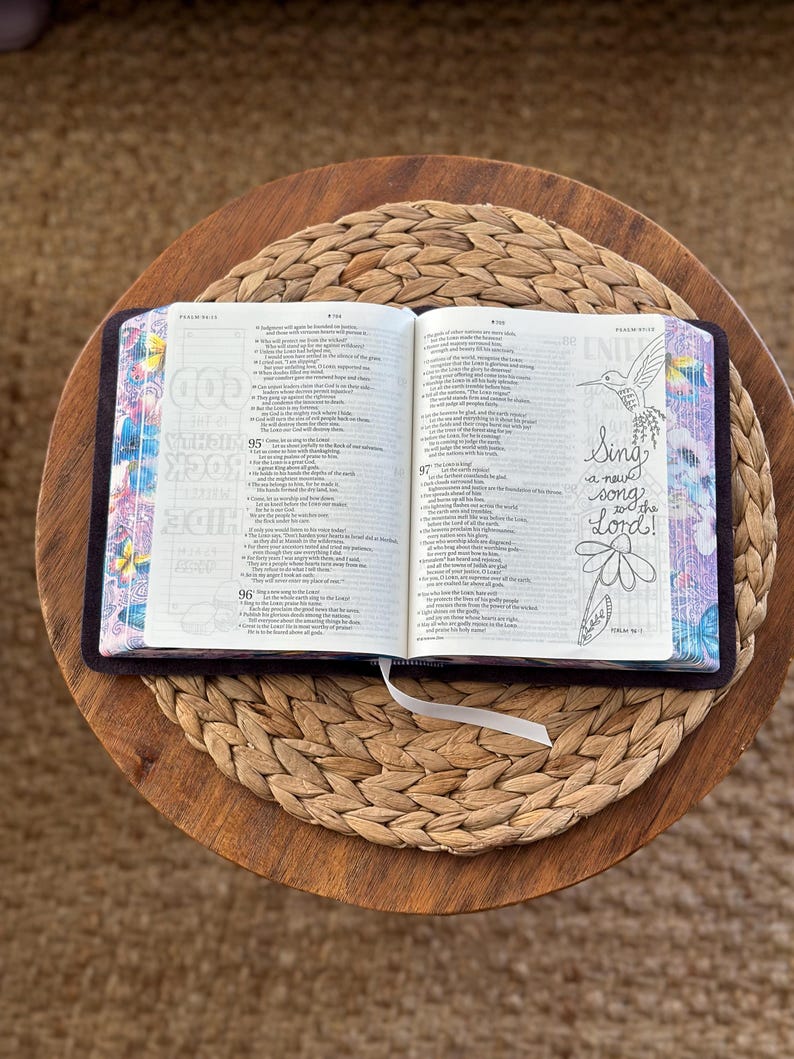 May include: An open Bible with dark covers and patterned page edges featuring butterflies and flowers. The right page displays a hand-drawn illustration of a hummingbird and a flower with the text, Sing a new song to the Lord!
