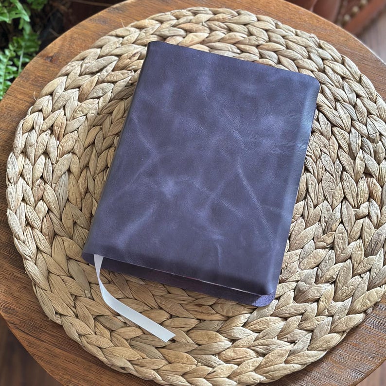 May include: A rectangular notebook with a dark purple leather cover featuring a smooth, matte texture and visible natural grain patterns. A thin, white ribbon bookmark extends from the bottom edge of the closed book, which is positioned centrally.